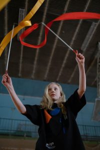 Child executing a ribbon gymnastics routine in an indoor sports hall, showcasing skill and concentration.